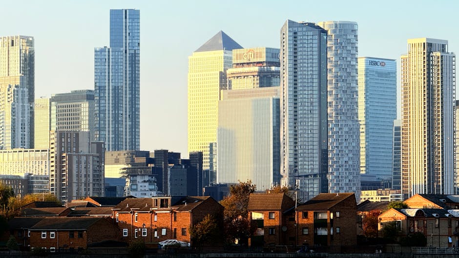 A view of a modern city skyline featuring tall glass office buildings and skyscrapers with reflective windows, situated behind a row of red-brick residential houses with pitched roofs. The residential area has small trees and parked cars in the foreground, with the buildings illuminated by natural daylight. This urban scene illustrates the contrast between domestic homes and commercial high-rises, relevant to house removals and moving services offered by Man and Van Poplar, especially in the context of home relocation and furniture transport within or to the densely built city environment.