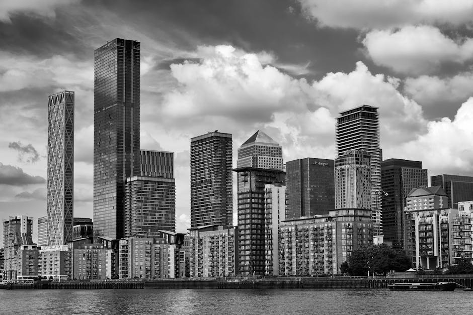 A black-and-white photograph of a city skyline featuring a cluster of modern high-rise buildings and skyscrapers with varied architectural styles, including glass facades and structural frameworks. In the foreground, there are mid-rise residential apartments situated along a waterfront, with visible balconies and windows. The scene is taken during daylight hours under partly cloudy skies, with some clouds casting shadows over the buildings. The background includes a partly cloudy sky, providing a contrast to the urban landscape. The image captures the essence of a bustling metropolitan area associated with house removals and furniture transport in a city like Canary Wharf, illustrating the type of environment that would be relevant for planning relocations or moving services, such as those offered by Man and Van Poplar.