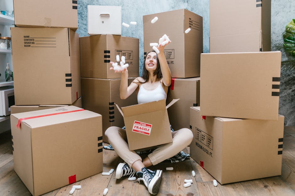 A young woman with long brown hair sitting cross-legged on a wooden floor inside a room filled with large cardboard moving boxes, some stacked on top of each other and sealed with packing tape. She is